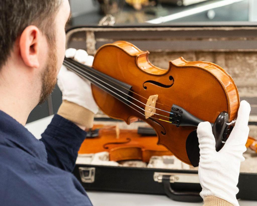 A man in a dark blue sweater carefully holds a shiny wooden violin. He wears white gloves to avoid damaging the violin as he lifts it from a black case, which is partially visible in the image. The background is blurred, but elements of an interior and other art objects can be discerned.