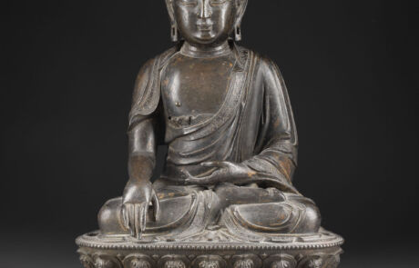 A detailed bronze statue of a seated Buddha on an elaborately decorated lotus throne. The Buddha wears a traditional robe and a crown, sitting in a meditation posture with his right arm raised and his hand resting flat on his knee. The background is uniformly dark, highlighting the statue.