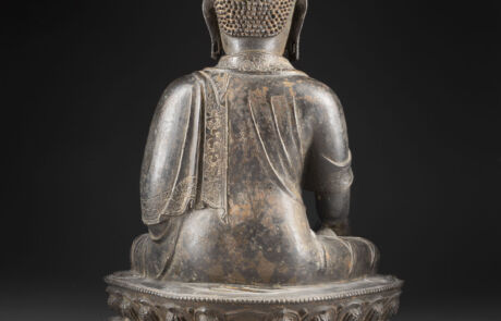 Rückansicht einer antiken Buddhastatue, sitzend auf einem reich verzierten Sockel. Die Statue zeigt detaillierte Falten in der Robe und feine Gravuren am Rücken und auf der Oberfläche des Sockels. Der Hintergrund ist dunkel, was die Statue hervorhebt.