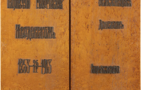 Zwei vertikal aufgestellte rechteckige Platten, möglicherweise aus Metall, mit einer rostigen orangen Oberfläche. Beide Platten sind mit erhabenen Buchstaben graviert, die schwer zu entziffern sind, eventuell Namen oder Titel in einer unbestimmten Schrift.