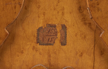 Nahaufnahme einer Holzoberfläche, möglicherweise Teil eines Kunstwerks oder Möbelstücks. Zwei alte, teils abgelöste Papieretiketten mit handschriftlichen Beschriftungen und Zahlen sind zu sehen, was auf eine frühere Auktion oder Katalogisierung hinweist.