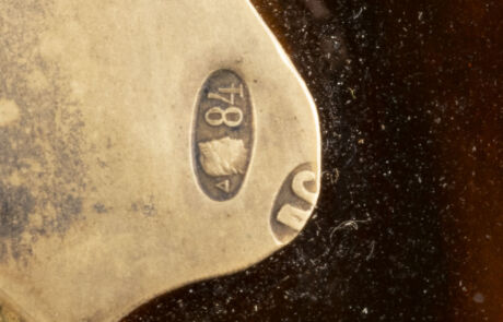 Nahaufnahme eines markanten Details eines Kunstobjekts, wahrscheinlich einem Teil eines Metallgegenstands. Im Fokus ist ein geprägtes, ovales Metallschild mit der Inschrift "18" und einem kleinen Symbol darunter, platziert auf einer leicht abgenutzten und mattierten Oberfläche. Der Hintergrund ist dunkel und unscharf, was das Metallschild hervorhebt.