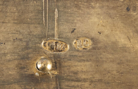 Nahaufnahme eines verwitterten Gemäldes zeigt schwer erkennbare, gravierte Details mit Jahrgang „1879“. Die Textur ist durch Alterung und Patina gezeichnet, was eine Mischung aus dunklen und helleren Brauntönen aufweist und das Kunstwerk historisch wertvoll erscheinen lässt.