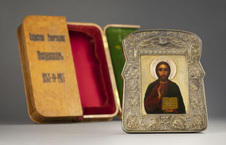 Porträt einer orthodoxen Ikone in silberner Relief-Umrahmung aufgestellt neben einer offenen, rechteckigen Box in Gold und Rot. Das Bildnis zeigt eine männliche Figur mit dunklem Bart und traditioneller geistlicher Gewandung, die ein Buch hält. Hintergrund ist neutral und unscharf, betont die Detailarbeit am Rahmen und der Ikone.