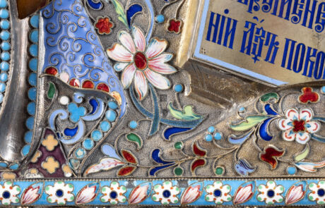 Nahaufnahme eines dekorativen Kunstobjekts, vermutlich Teil eines Auktionssorts vom Kunstauktionshaus Hargesheimer. Das Bild zeigt ein detailreiches, farbenfrohes Muster mit floralen Motiven und eingebetteten türkisen und blauen Steinen. Die Arbeit umfasst feine Metallarbeiten und farbige Emaillen.