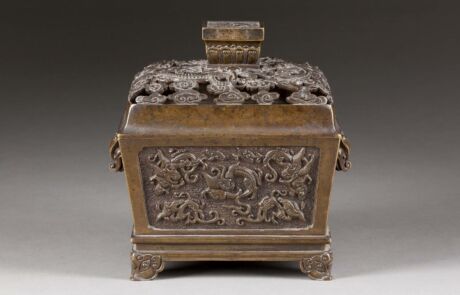 An antique, rectangular metal container on a small pedestal, probably made of bronze, with complex, engraved decorations on all sides. The lid of the container is crowned with finely crafted floral designs, terminating in a prominent, rectangular handle.