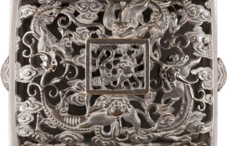 Detailaufnahme eines Kunstobjekts mit aufwendigen Silberschnitzereien, darstellend verschlungene, mythologisch anmutende Tiere und filigrane Muster. Das Zentrum schmückt ein quadratischer Rahmen mit einem komplexen, gravierten Motiv. Hintergrund und Materialien wirken antik, typisch für historische Wertstücke.