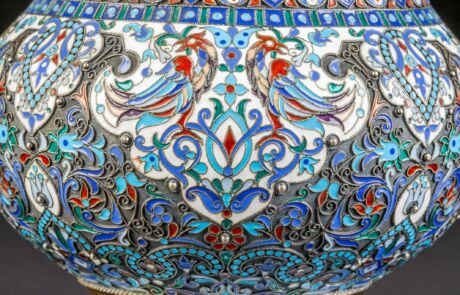 Detailaufnahme eines kunstvoll verzierten Objekts, vermutlich eine Vase, aus einem Auktionshaus. Das Dekor zeigt intensive blaue, rote und grüne Farben mit floralen und geometrischen Mustern, die von goldenen Linien umrahmt sind. Es besticht durch seine detailreichen Ornamente und präzise Handwerkskunst.