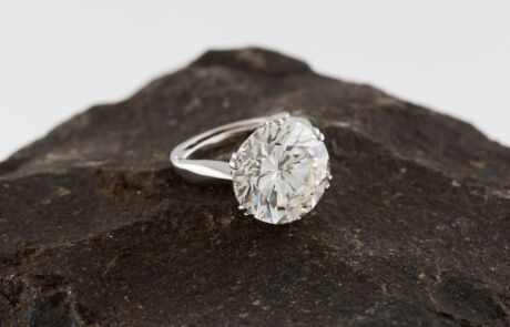 A lustrous diamond ring set on a dark, rough rock, featuring a large, brightly sparkling solitaire diamond in a silver setting. The diamond is round-cut, and the metal of the ring shines with its polish.