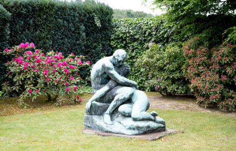 Patinierte Bronzeskulptur im Garten, die eine nachdenkliche, sitzende menschliche Figur darstellt, umgeben von grünen Sträuchern und rosa blühenden Pflanzen. Die Skulptur zeigt detaillierte, ausdrucksstarke Züge und glatte Oberflächen und ist im Freien aufgestellt.