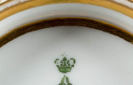 Nahaufnahme eines antiken Porzellantellers mit grüner Krone auf weißem Hintergrund, umrandet von Goldverzierungen.