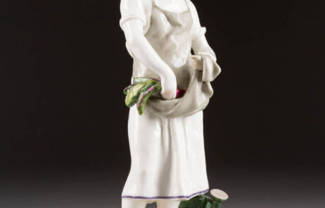 Antike Porzellanskulptur einer stehenden Figur mit gesenktem Blick und Spargelbündel in der Hand, präsentiert bei einer Kunstauktion.