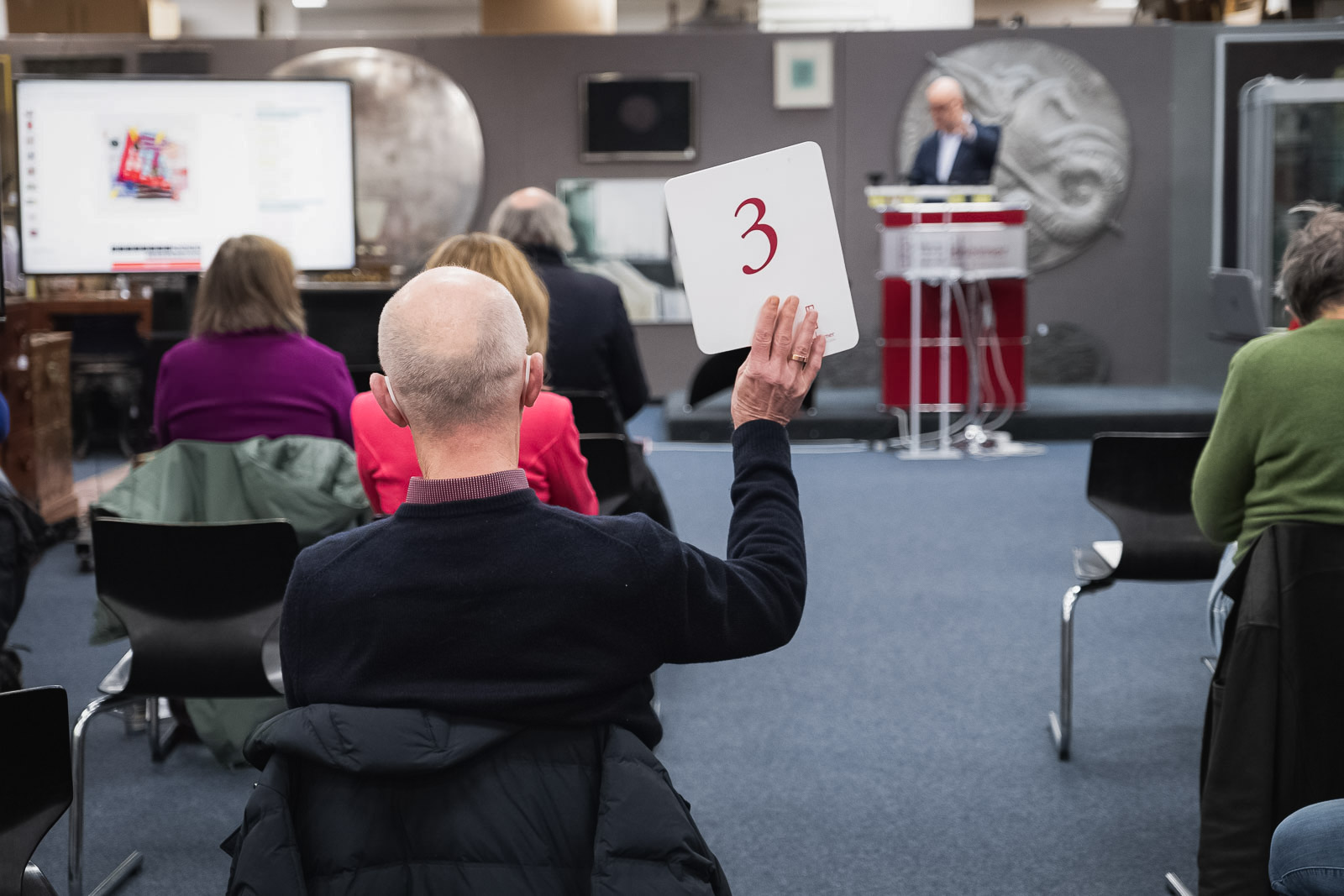 Eine Auktionsszene mit einem Mann, der eine Nummer 3 hochhält, im Hintergrund ein Auktionator und andere Bieterinnen und Bieter.