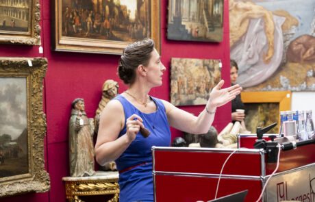 Frau in blauem Kleid leitet Auktion in einem Raum mit roten Wänden, umgeben von verschiedenen Kunstwerken, darunter Gemälde und Skulpturen.