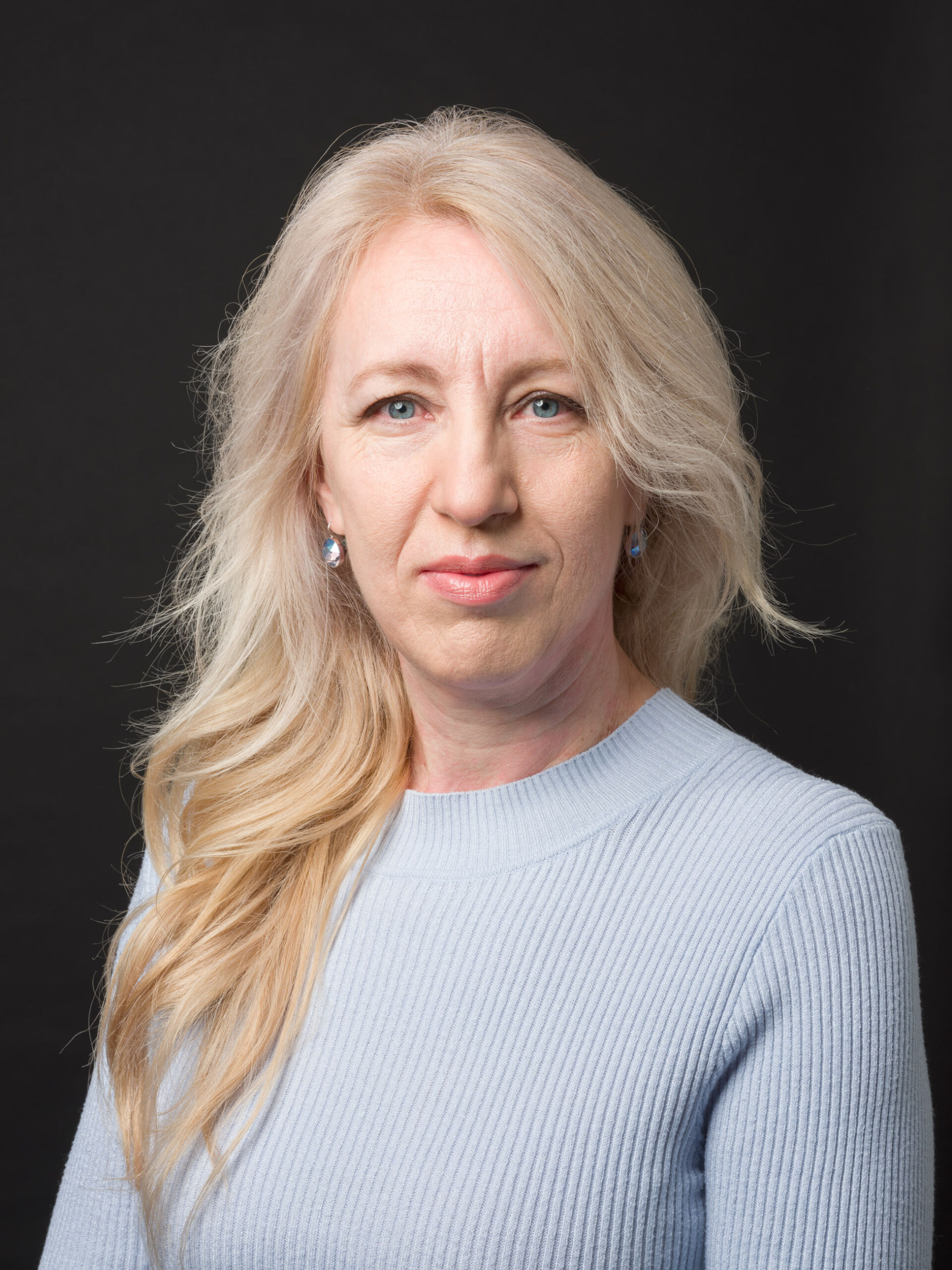 Porträt einer Frau mittleren Alters mit langen, welligen blonden Haaren und hellblauer Kleidung. Die Frau schaut direkt in die Kamera, vor einem einfarbigen dunklen Hintergrund. Sie trägt Ohrringe, die zu ihrer Kleidung passen, und hat ein subtiles Lächeln.