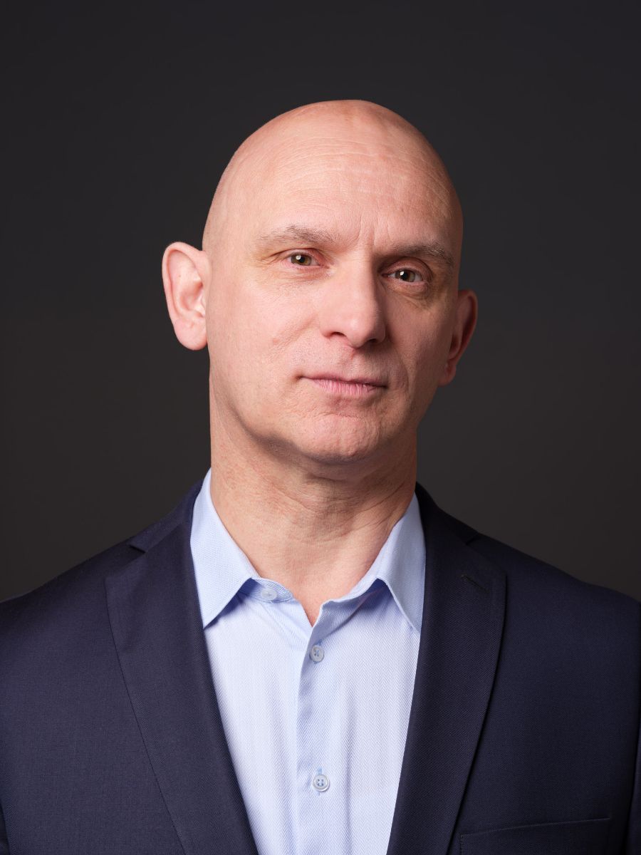 Porträt eines Mannes in dunklem Anzug und hellblauem Hemd vor dunklem Hintergrund.