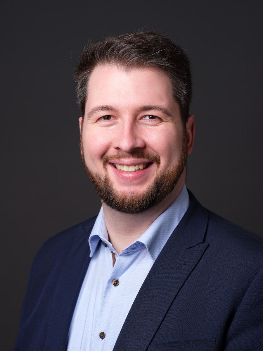 Porträtfoto eines lächelnden Mannes mit Bart, blauem Hemd und dunklem Sakko vor einem grauen Hintergrund.