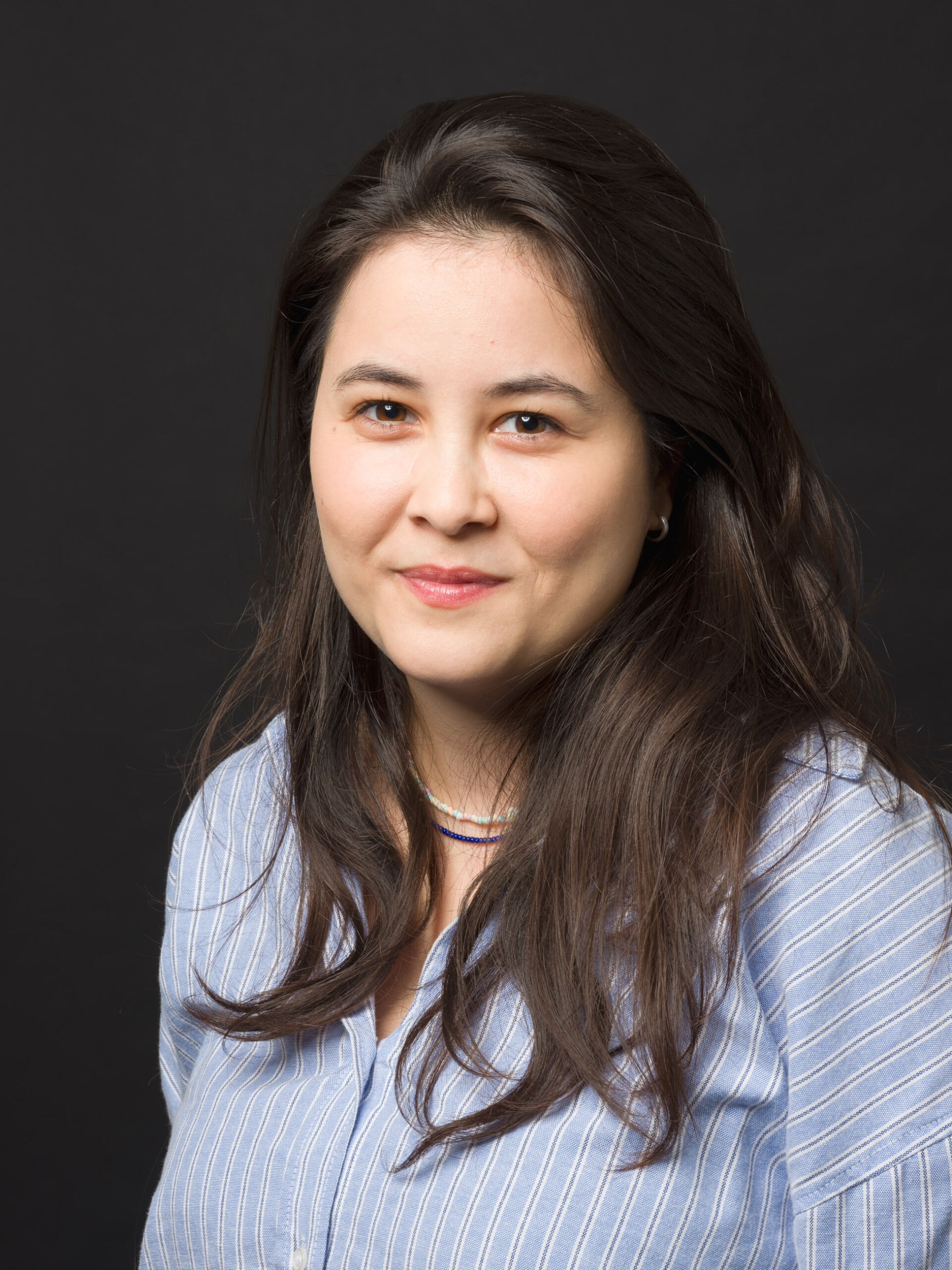 Porträt einer Frau mit asiatischen Merkmalen, die freundlich in die Kamera lächelt. Sie trägt ein hellblaues gestreiftes Hemd und hat langes dunkles Haar, das offen über ihre Schultern fällt. Der Hintergrund des Bildes ist einheitlich dunkelgrau.
