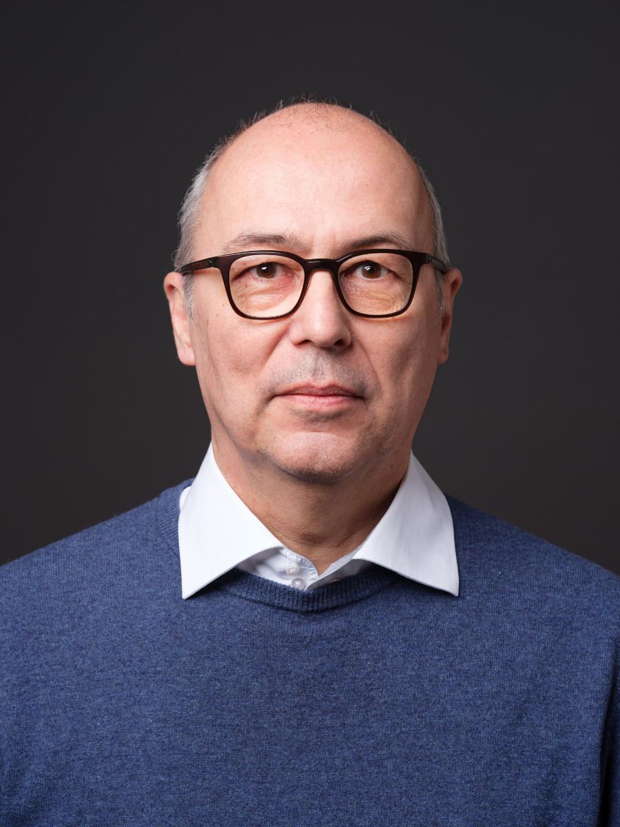 Porträt eines Mannes mit Brille, der einen blauen Pullover und darunter ein weißes Hemd trägt, vor einem dunkelgrauen Hintergrund.