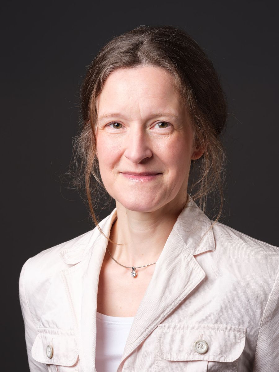 Portrait einer lächelnden mittelalten Frau mit zurückgebundenen braunen Haaren. Sie trägt eine helle Khakijacke, ein weißes Top und eine Halskette mit einem kleinen Anhänger. Der Hintergrund des Bildes ist einheitlich dunkelgrau.