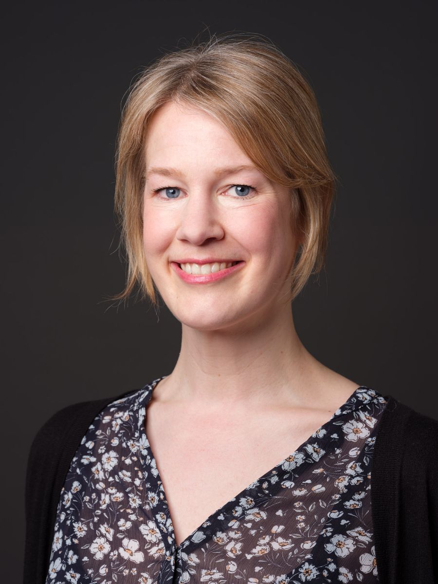 Porträtfoto einer lächelnden Frau mit blauem Blumenkleid vor dunklem Hintergrund.