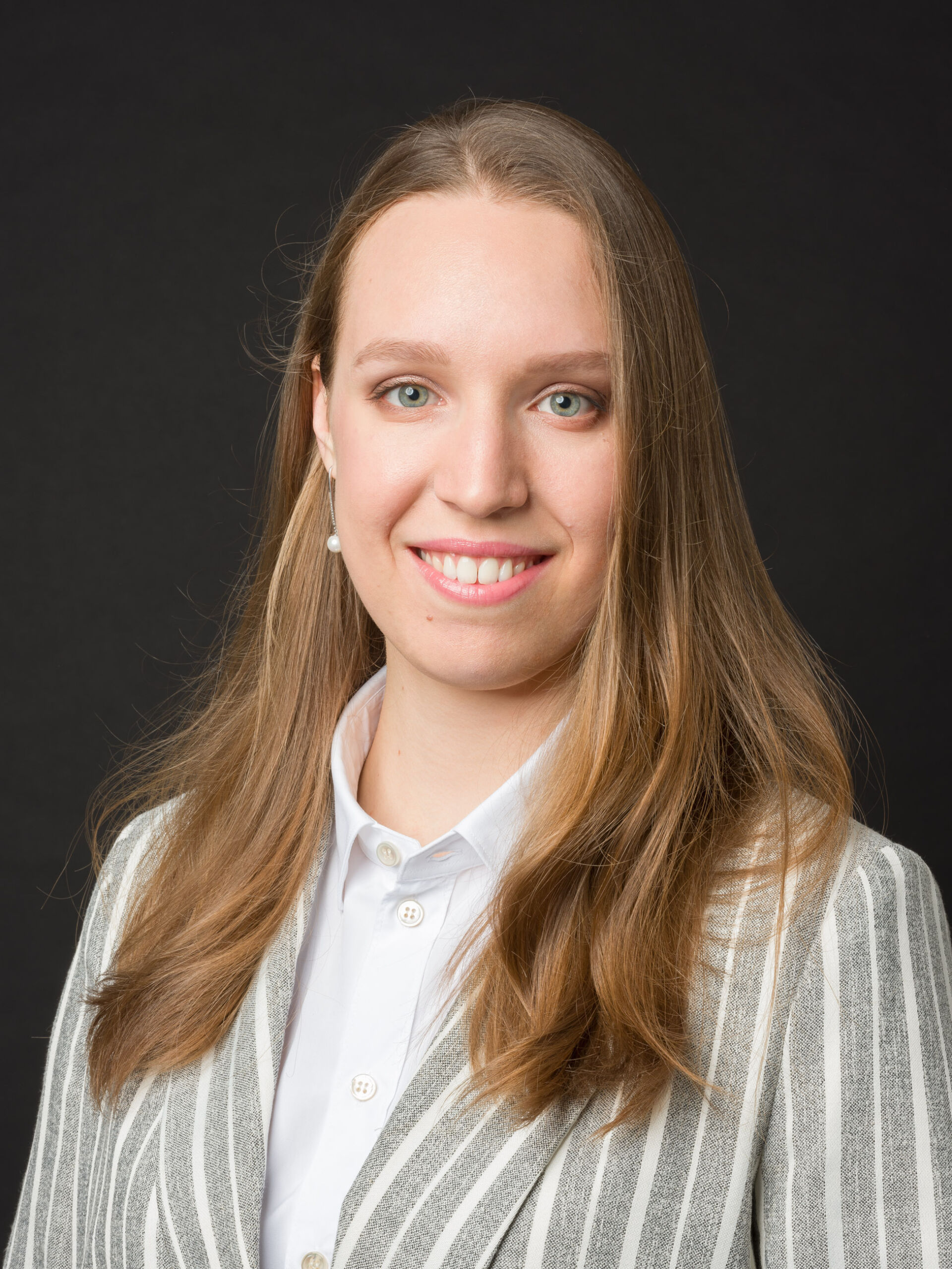 Porträtfotografie einer jungen Frau mit langen, hellbraunen Haaren, die vor einem dunklen Hintergrund posiert. Sie trägt ein weißes Hemd unter einem grauen gestreiften Blazer und perlmuttfarbene Ohrringstuds. Sie lächelt freundlich und hat auffallende, klare blaue Augen. Die Beleuchtung betont ihre Gesichtszüge.