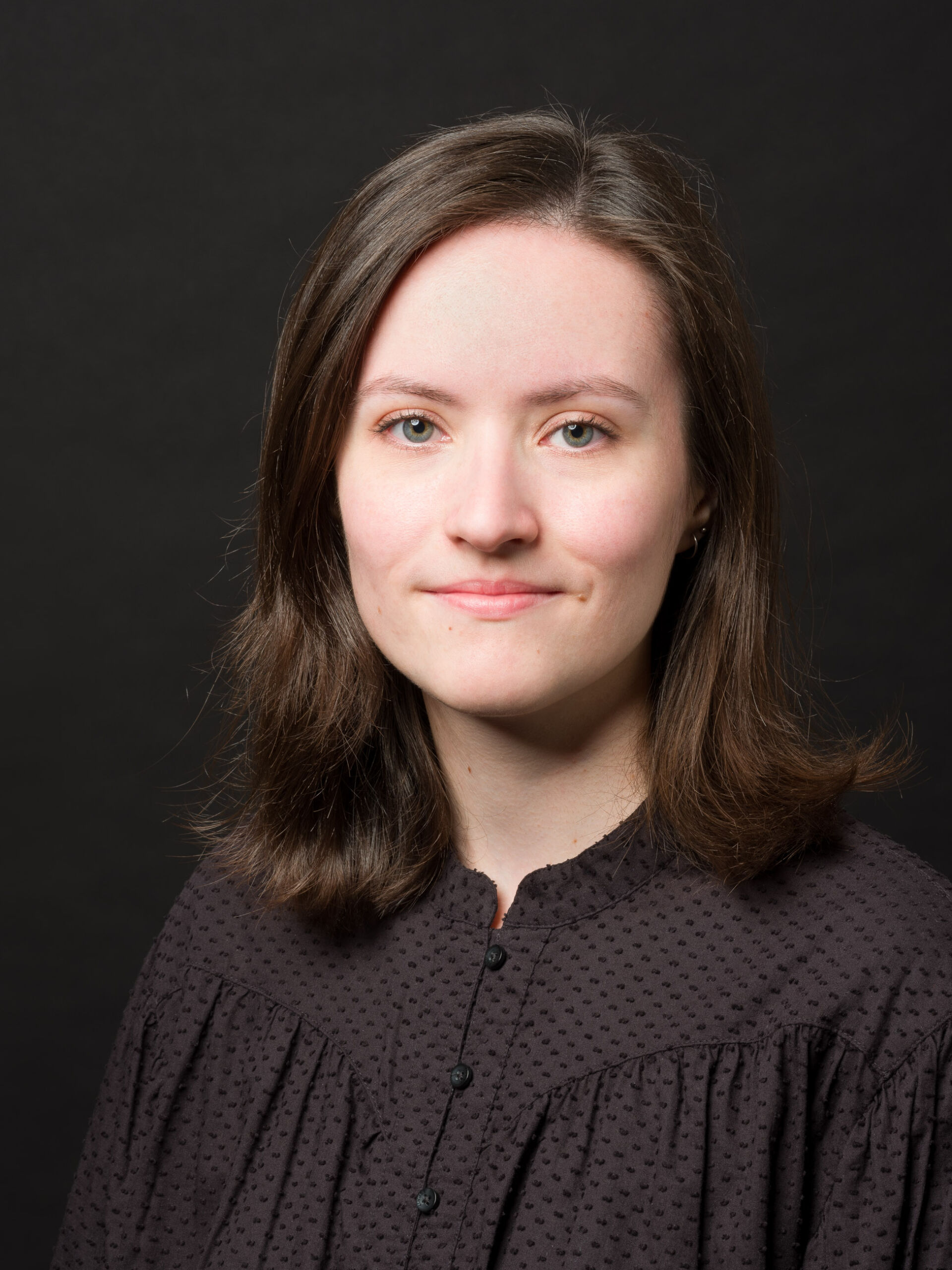 Porträtfoto einer jungen Frau mit schulterlangem braunem Haar, die direkt in die Kamera blickt. Sie trägt eine dunkle, gepunktete Bluse mit kleinen, runden Knöpfen. Der Hintergrund ist einheitlich dunkelgrau. Das Bild wird von weichem, natürlich wirkendem Licht erhellt, das sanfte Schatten auf ihr Gesicht wirft und ihre Grübchen leicht hervorhebt.
