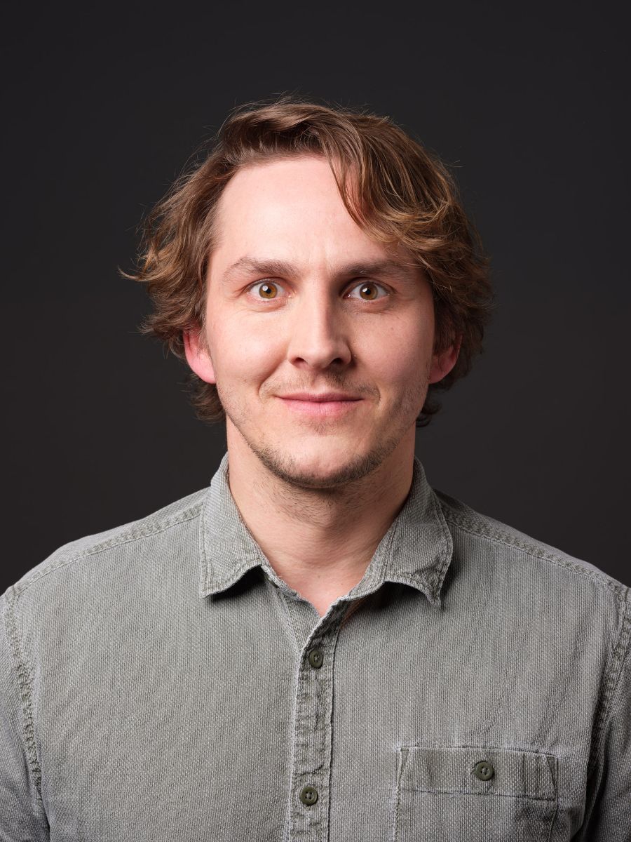 Porträt eines lächelnden Mannes mit mittellangem, gewelltem braunen Haar und leichten Gesichtszügen. Er trägt ein graues Hemd mit aufgeknöpftem Kragen und blickt direkt in die Kamera. Der Hintergrund ist einheitlich dunkelgrau, fokussierend die Aufmerksamkeit auf das freundliche Gesicht. Er hat hellbraune Augen.