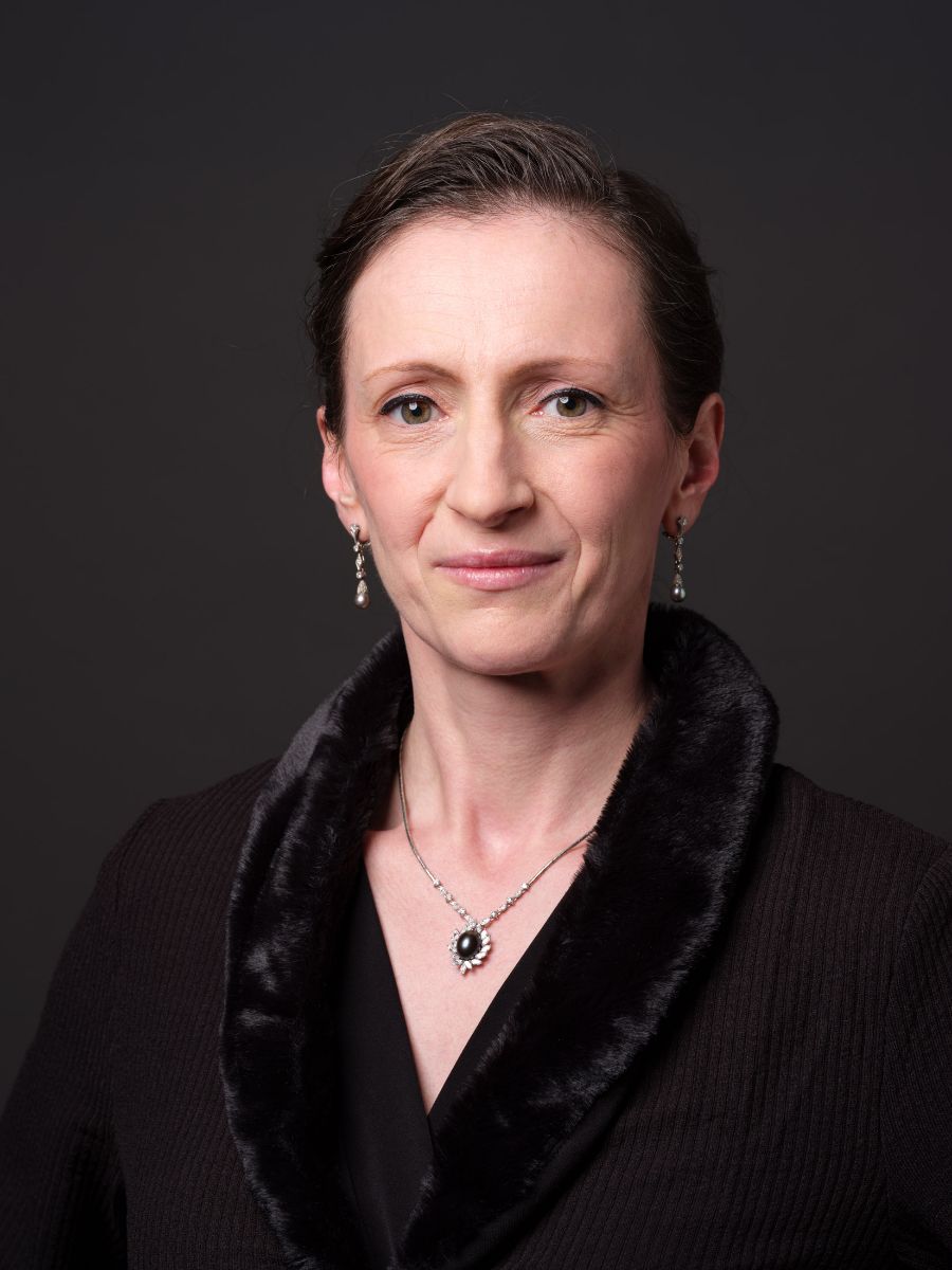 Porträt einer Frau (Susanne Hargesheimer)mit kurzem, dunklem Haar und samtigem Kleid. Sie trägt diamantbesetzte Ohrringe und eine Halskette mit schwarzem Edelstein. Der Hintergrund ist dunkel.