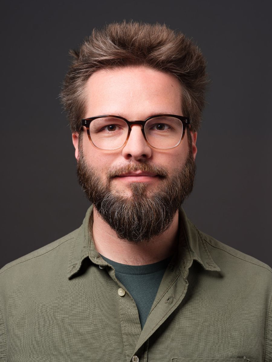 Porträtfoto eines Mannes mit Brille, Bart, gekleidet in ein grünes Hemd und ein blaugrünes T-Shirt darunter.