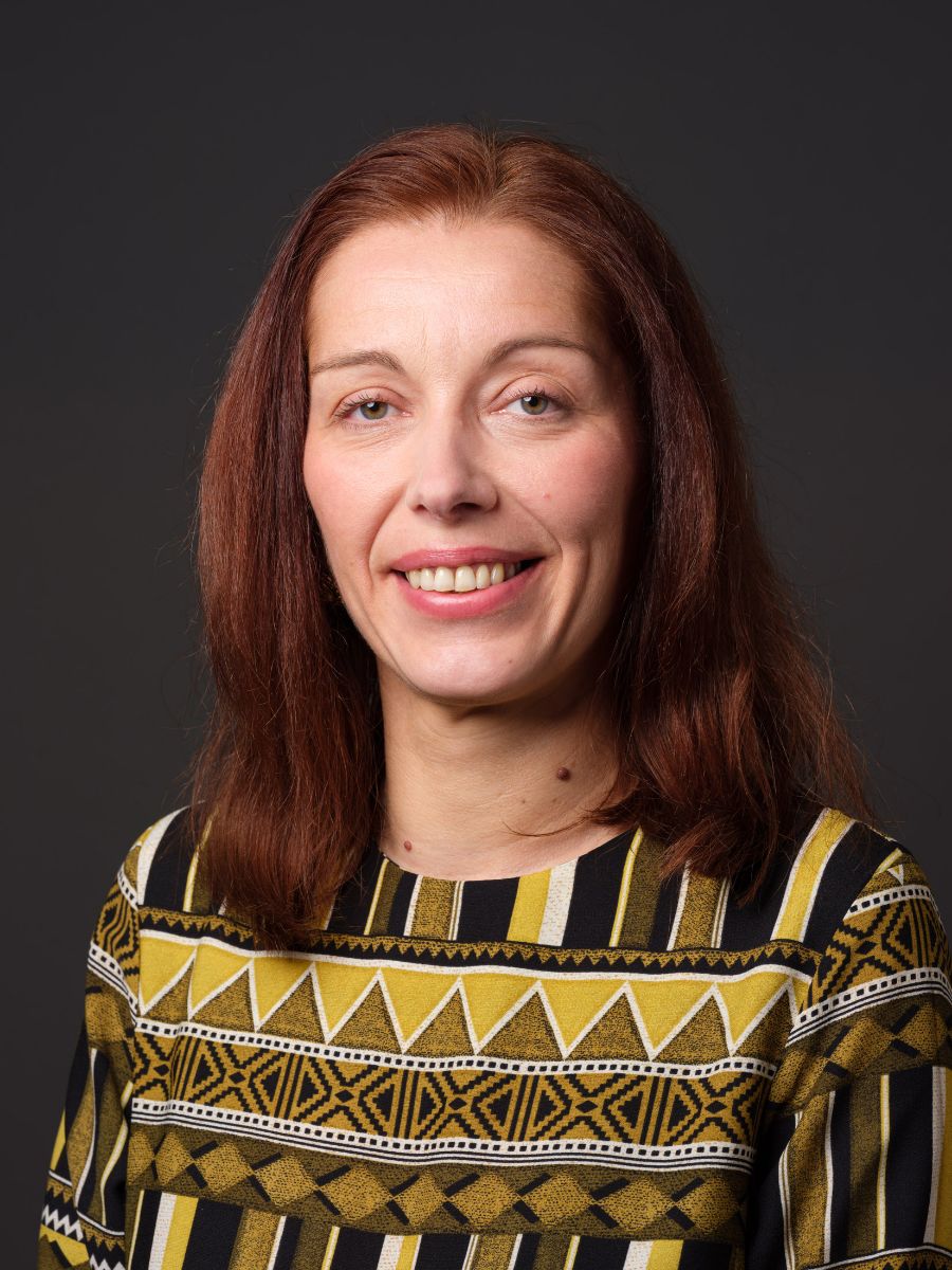 Porträtfoto einer lächelnden Frau mit langen roten Haaren, die ein gemustertes, mehrfarbiges Oberteil trägt, vor dunklem Hintergrund.