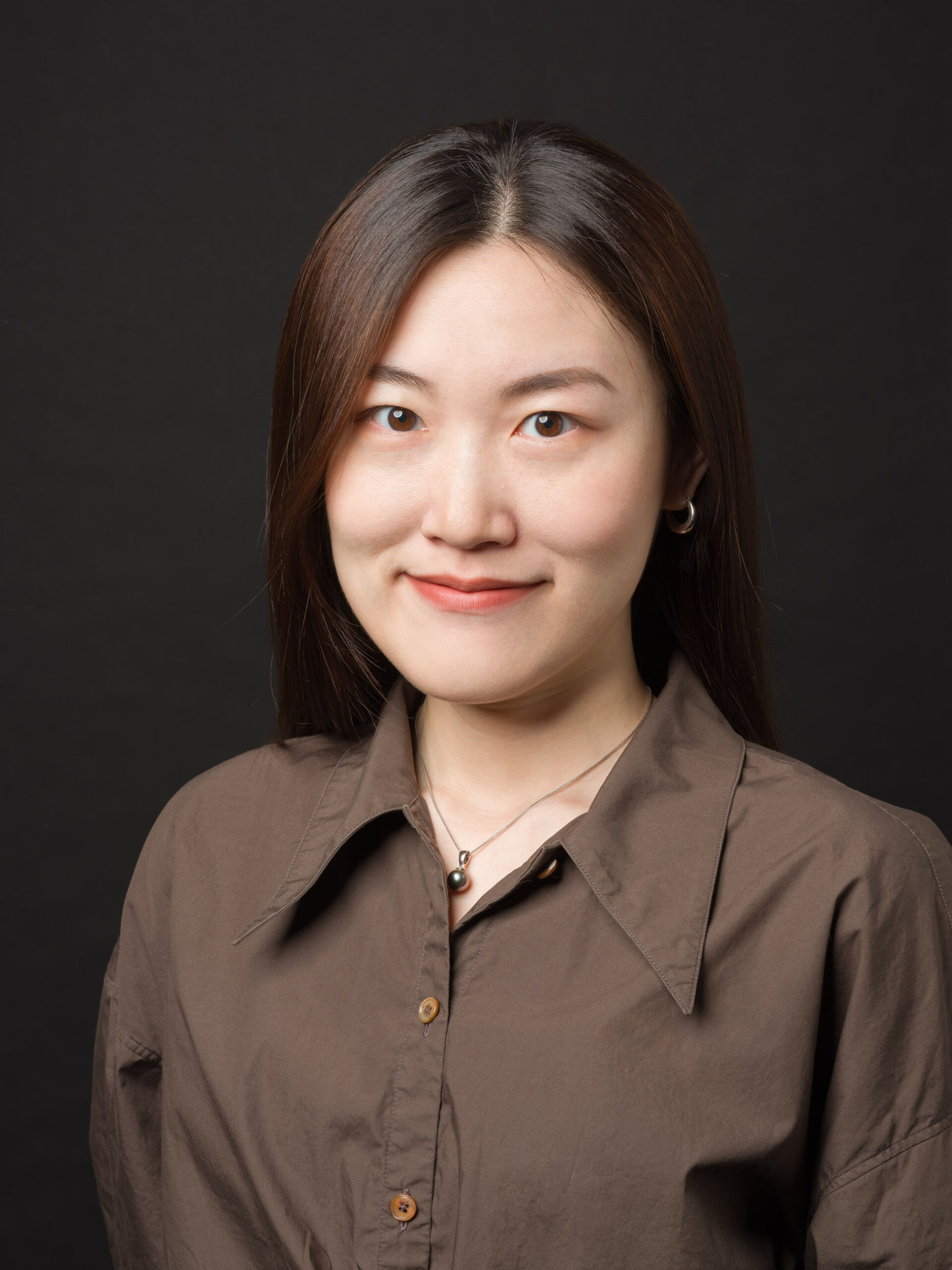 Porträtfoto einer jungen asiatischen Frau auf schwarzem Hintergrund. Sie trägt eine braune Bluse und eine dezente Halskette mit einer dunklen Perle. Ihr mittellanges dunkles Haar fällt glatt über die Schultern. Sie lächelt sanft in die Kamera, und ihre Augen haben einen freundlichen Ausdruck.