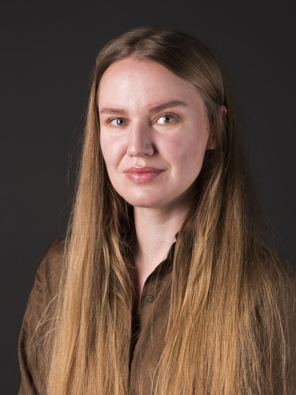 Porträtfoto einer jungen Frau mit langen, glatten, blonden Haaren und einem braunen Hemd vor einem dunklen Hintergrund. Sie blickt direkt in die Kamera, ihr Gesichtsausdruck ist neutral, und das Licht betont ihre Gesichtszüge sanft.