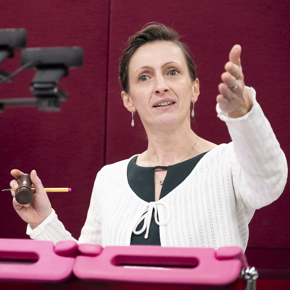 Frau in weißer Bluse steht vor einem roten Hintergrund und hält einen Auktionshammer in der Hand, während sie mit der anderen Hand deutend gestikuliert.