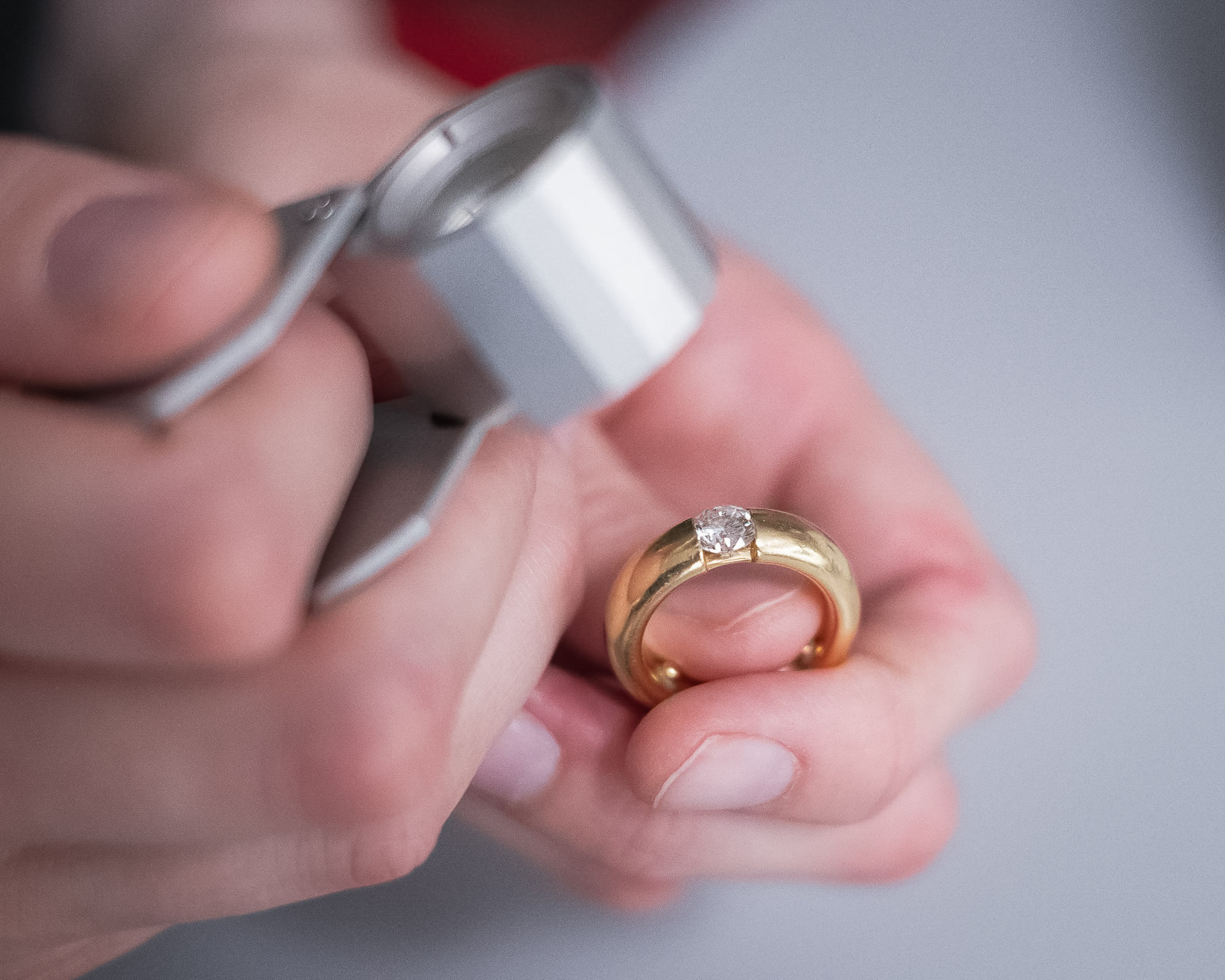 Nahaufnahme zweier Hände, die mittels einer Lupe einen goldenen Ring mit großem Diamanten begutachten.