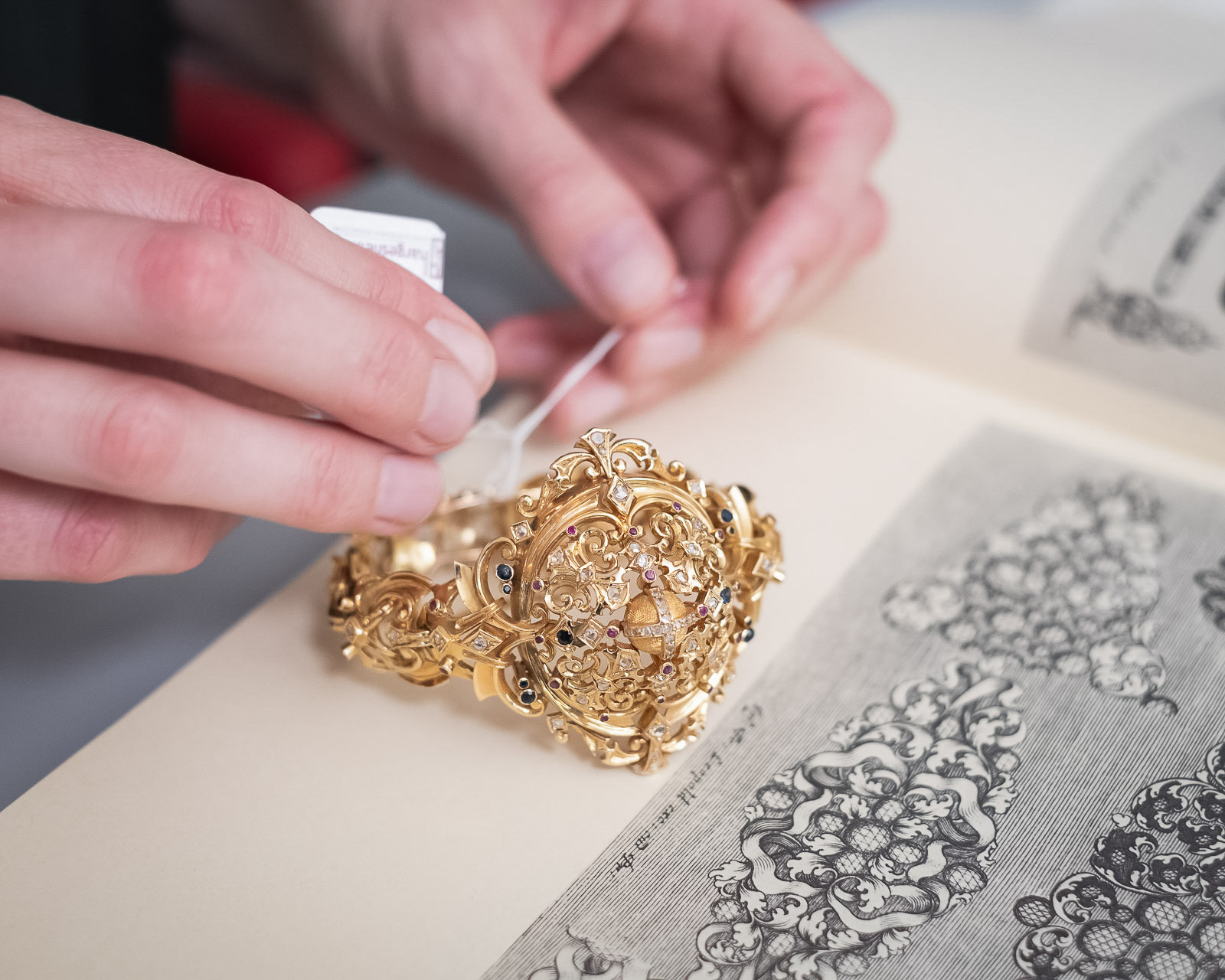 Detailaufnahme einer Hand, die ein fein gearbeitetes goldenes Schmuckstück über einem Buch mit klassischen Drucken prüft.