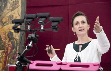 Auktionsleiterin dirigiert lebhaft eine Kunstauktion, umgeben von historischen Gemälden und moderner Ausrüstung.