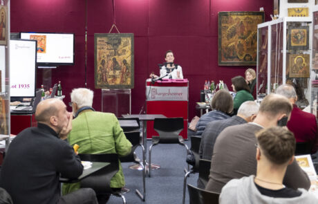 Auktionsszene in einem Raum gefüllt mit Ikonen, eine Frau bei der Arbeit, weitere Personen im Hintergrund, moderne Technologie für die Übertragung.