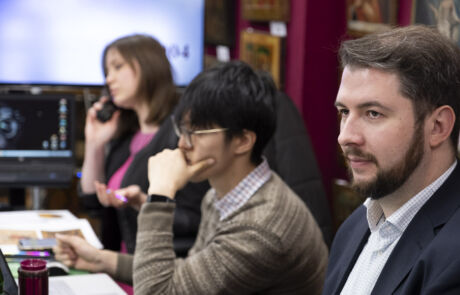 Drei Personen bei einer Kunstauktion sitzen vor Mikrofonen; eine Frau und zwei Männer analysieren Kunstwerke in einem Innenraum mit Gemälden im Hintergrund.