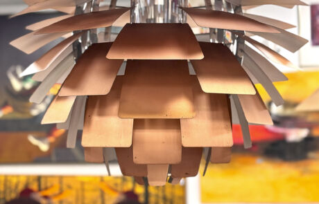 Kupferfarbene Hängelampe im modernen Design, präsentiert in einem Auktionssaal mit unscharfen Kunstwerken im Hintergrund.