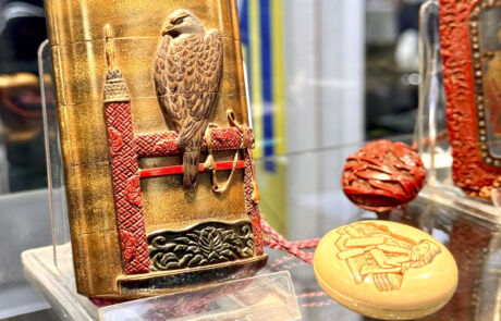 Asiatisches Inrō mit Relief eines Falken auf einem roten und goldenen Thron, flankiert von einem geschnitzten roten und goldenen Skulpturobjekt und einer reliefverzierten elfenbeinfarbenen Dose.