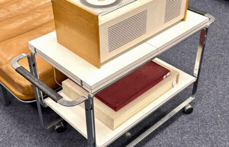 Vintage Braun-Musikanlage "Schneewittchensarg" auf mobilem Rolltisch neben einem Ledersessel.