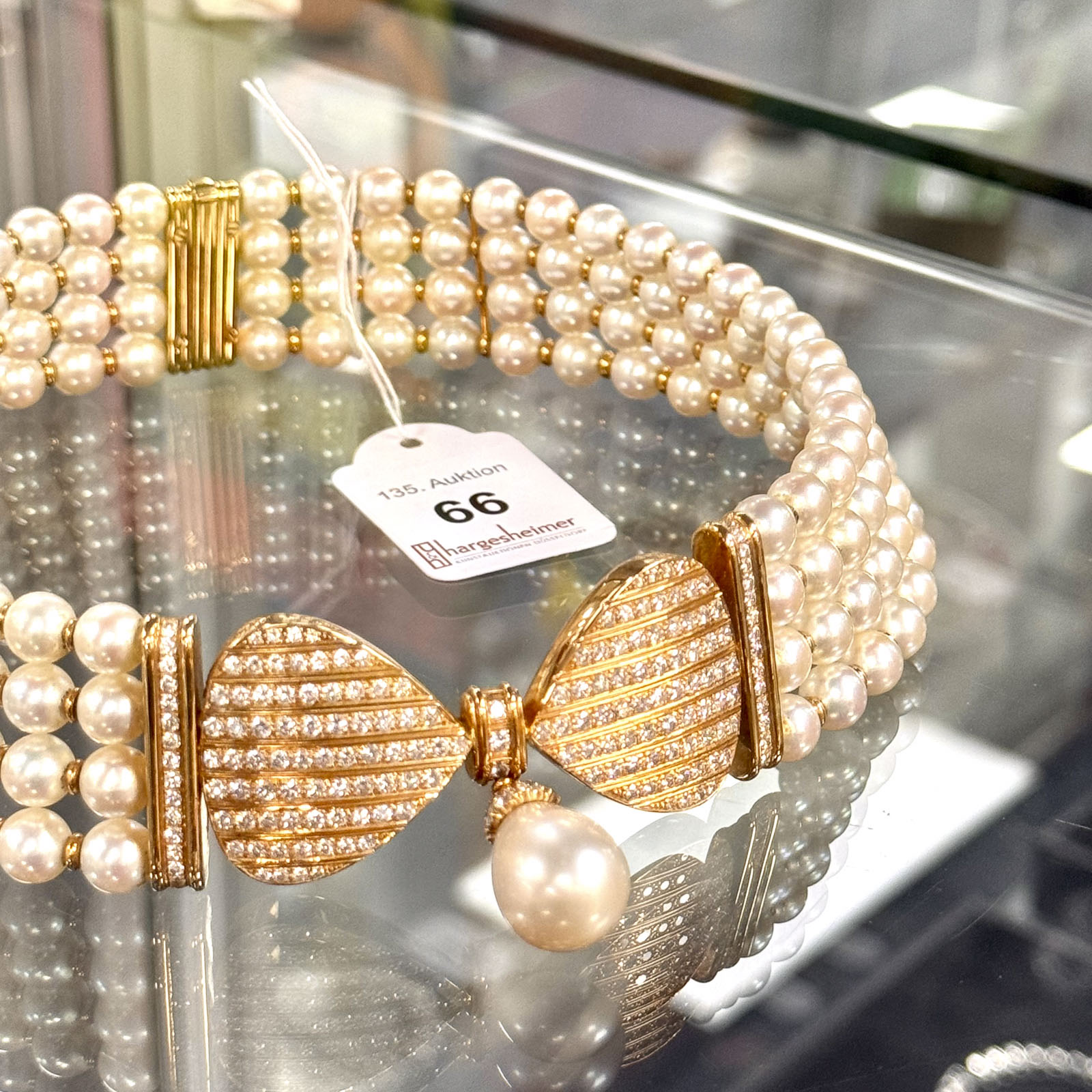 Perlenschmuck in einer Vitrine, bestehend aus mehrreihigen Perlencolliers und einem zentralen goldfarbenen Verschluss, besetzt mit zahlreichen kleinen Diamanten und einer großen Perle. An einem weißen Etikett ist die Nummer "66" und "135. Auktion" vermerkt.