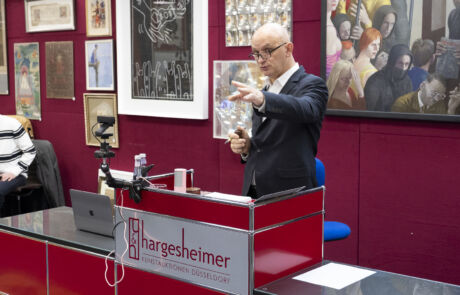 Auktionator präsentiert ein Glasobjekt bei einer Kunstauktion bei Hargesheimer; rote Wände mit hängenden Bildern im Hintergrund.