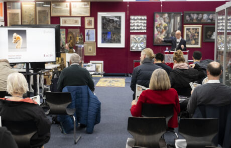 Auktionssaal mit Bietern bei Hargesheimer, Auktionator präsentiert historisches Kunstwerk auf Bildschirm.