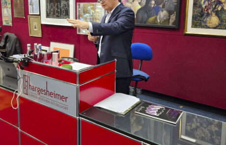 Auktionator leitet Kunstauktion bei Hargesheimer, umgeben von historischen Kunstwerken und Sammlerstücken.