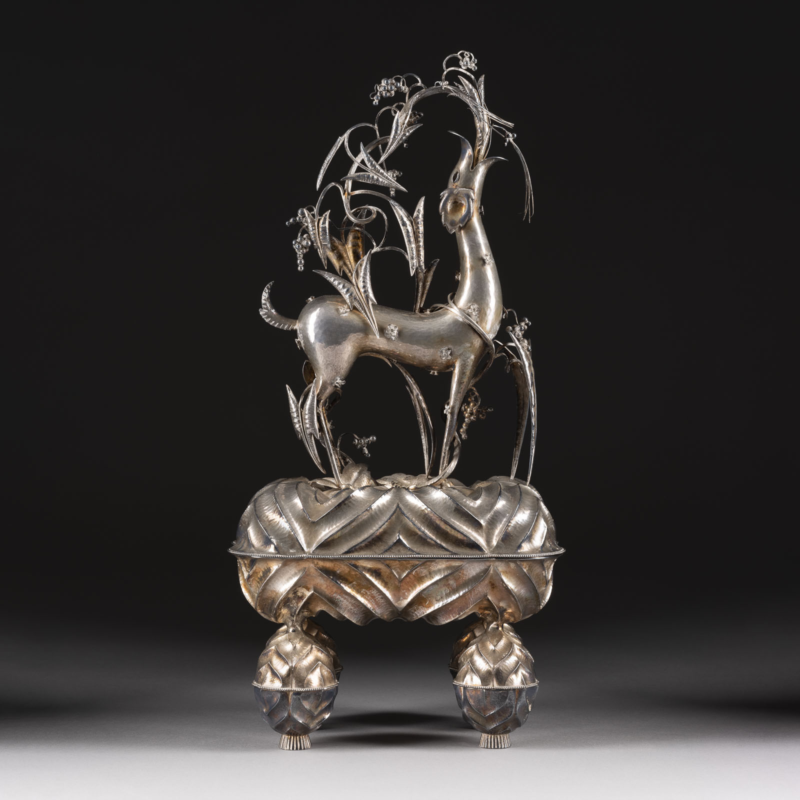 Silberne Skulptur eines springenden Fabeltiers auf einem reich verzierten Podest, umgeben von filigranen Pflanzen und Blumen, vor neutralem Hintergrund.