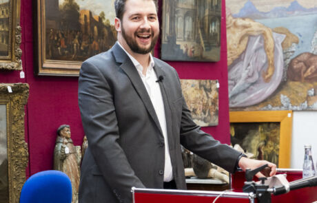 Auktionator leitet Kunstauktion bei Hargesheimer, umgeben von wertvollen Gemälden und Skulpturen.