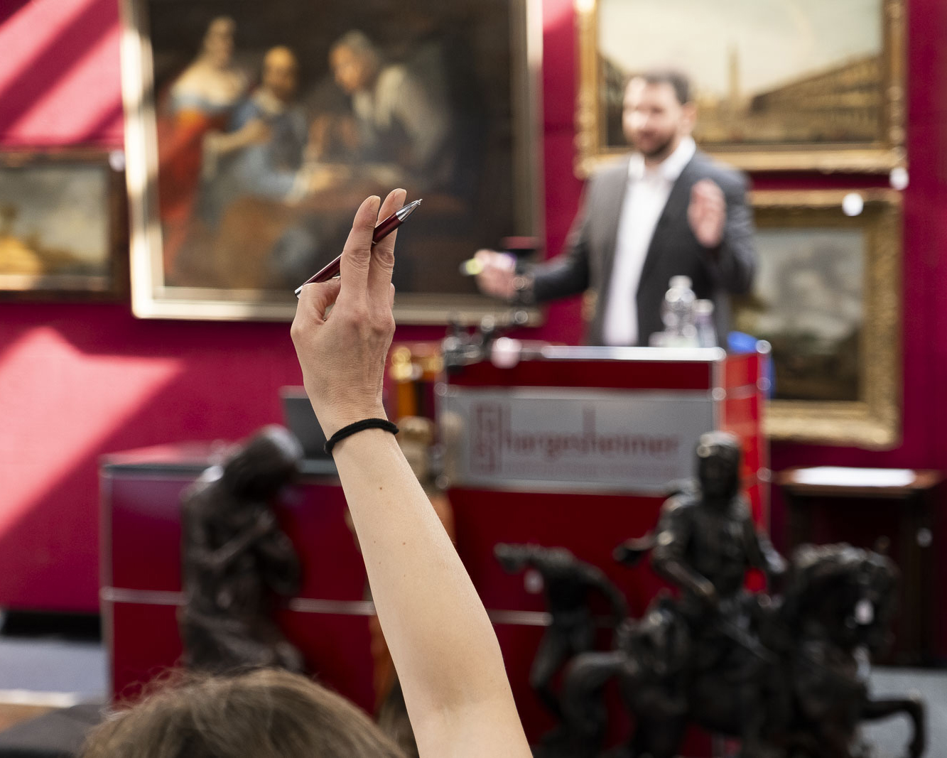 Eine Kunstauktion bei Hargesheimer in Düsseldorf, mit einer Person, die ein Gebot abgibt, während der Auktionator im Hintergrund steht. Im Raum sind verschiedene klassische Gemälde und Skulpturen zu sehen.