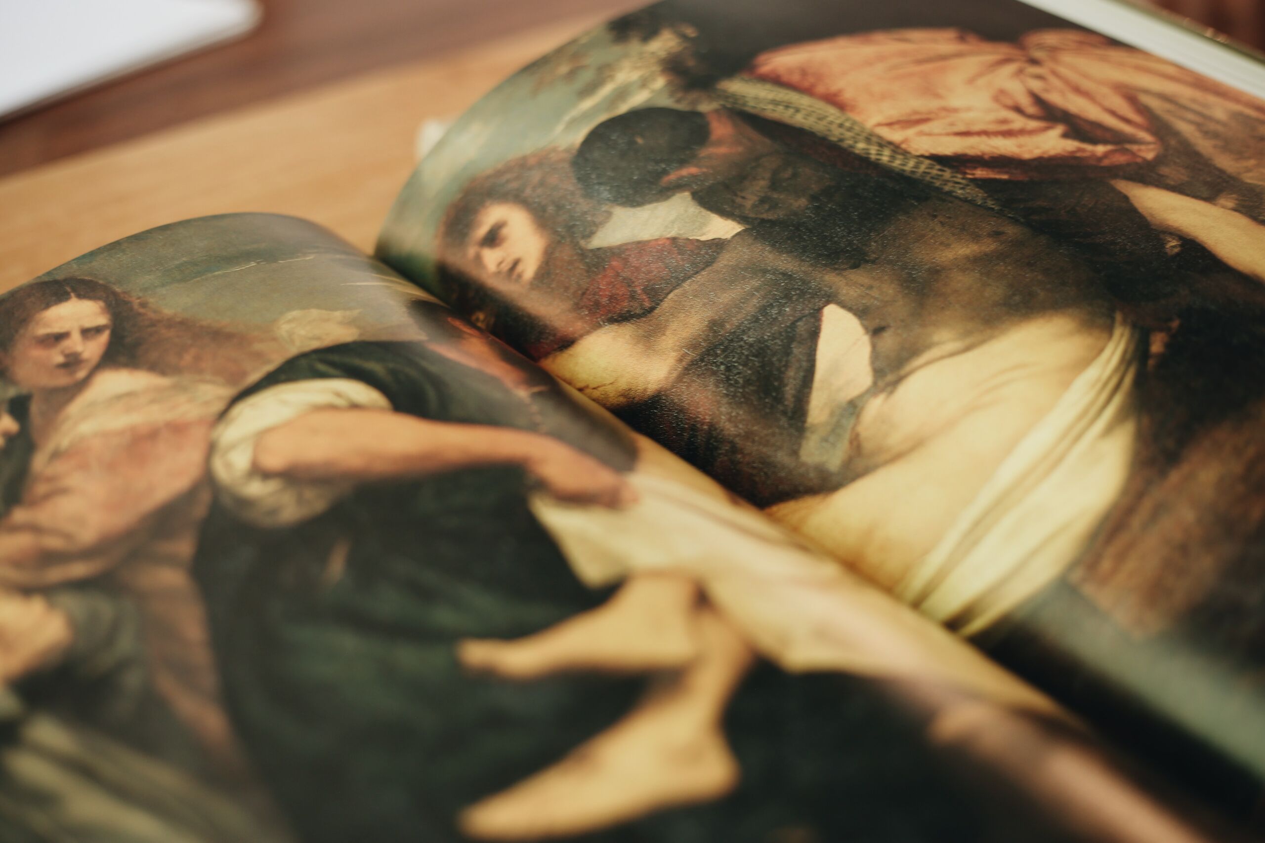 Nahaufnahme eines aufgeschlagenen Kunstbuches, das zwei klassische Gemälde zeigt. Das linke Bild präsentiert drei lebensnah dargestellte Figuren, von denen eine den Blick direkt zum Betrachter richtet. Das rechte Bild zeigt eine liegende, teils verdeckte Figur in gedämpften Farbtönen.